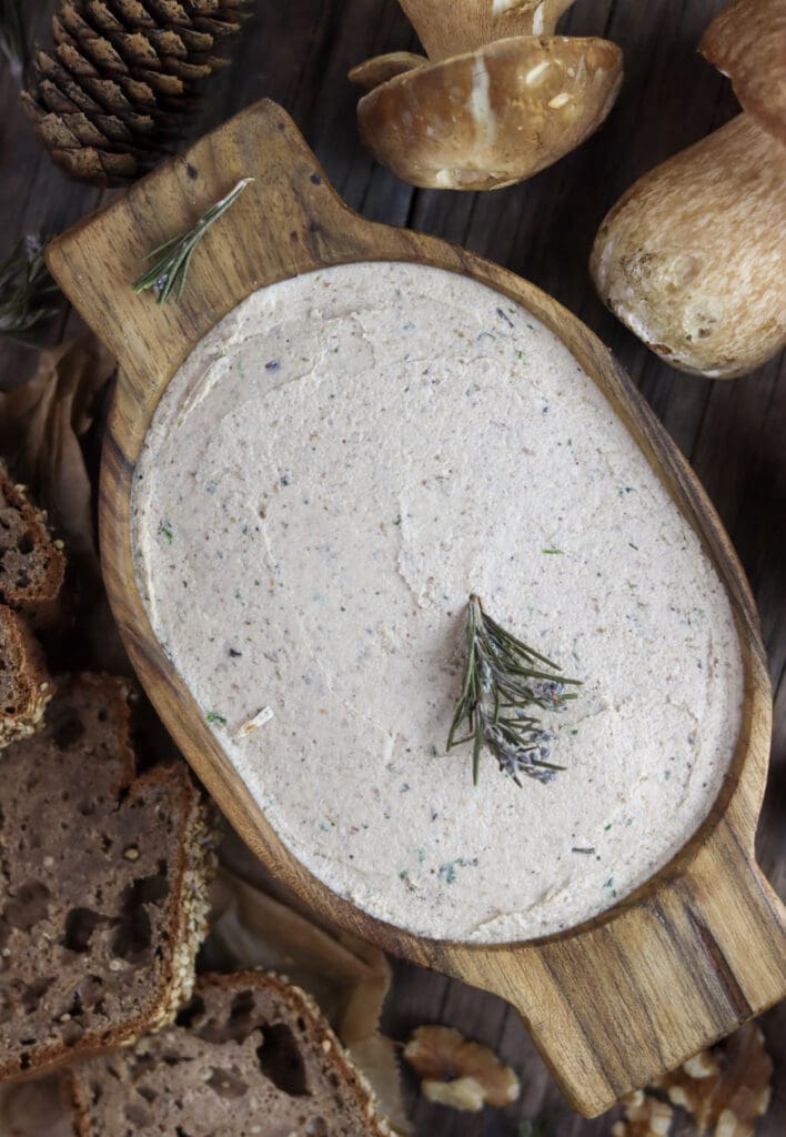 A wooden bowl of porcini mushroom walnut pate, decorated with rosemary with homemade bread and porcini mushrooms in the background.